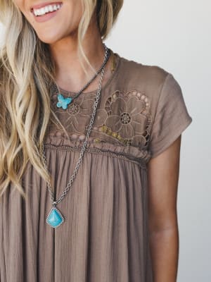 A close-up detail shot of a mocha gauze dress with floral crochet yoke and gathered skirt, styled with layered turquoise stone necklaces — the kind of outfit that feels like it was made for a slow summer afternoon.