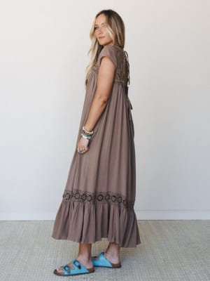 A flowy mocha maxi dress with crochet floral trim at the hem and lace-up back detail, styled with turquoise sandals and a stack of boho bracelets — the kind of dress that looks equally at home at a farmers market or a backyard wedding.