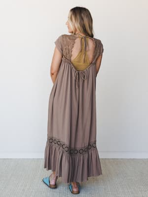 Shot from behind, this flowy mocha maxi dress does the quiet work of an outfit that knows it's special — open back with crochet floral cutout detail, a mustard yellow crochet inset panel, and a tiered ruffle hem with matching floral lace trim, worn w