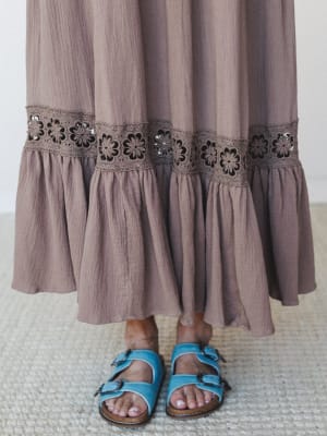 A close-up of a flowy maxi skirt hem in mocha gauze, detailed with a wide floral crochet trim band and a ruffled bottom tier, paired with blue buckle sandals on a woven rug.