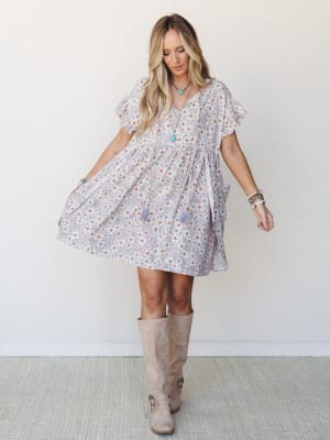 A flowy lavender floral mini dress with flutter sleeves and a babydoll silhouette, styled with tall taupe suede boots and layered turquoise jewelry — the kind of outfit that looks equally at home at a farmers market or a backyard wedding.