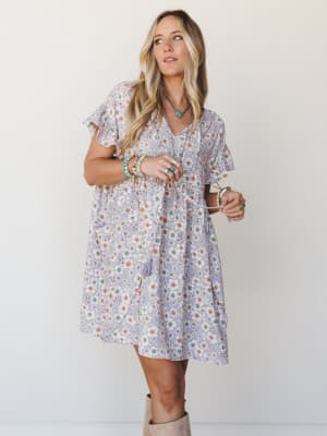 A flowy lavender floral mini dress with flutter sleeves and a relaxed babydoll silhouette, styled with turquoise jewelry and tan boots for an easy boho-everyday look.