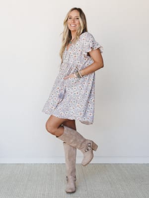 A flowy lavender floral mini dress with flutter sleeves and a babydoll silhouette, styled with tall suede boots and layered boho jewelry — the kind of outfit that looks equally at home at a farmers market or a fall birthday dinner.