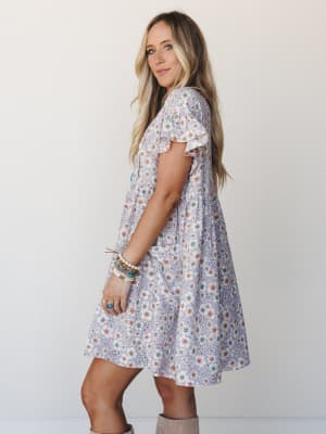 A flowy lavender floral mini dress with flutter sleeves and a relaxed babydoll silhouette, styled with stacked boho bracelets and taupe knee-high boots against a clean white studio backdrop — effortless and a little dreamy.