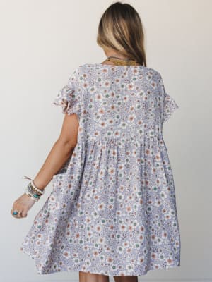 A breezy floral mini dress in soft lavender shot from the back, styled with layered boho jewelry and stacked bracelets — the kind of dress that makes a Tuesday feel like something worth dressing for.