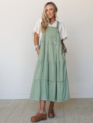 A tiered gingham midi overall dress in sage green layered over a white ruffle-sleeve blouse, styled with wood-platform clogs and stacked beaded bracelets — the kind of outfit that makes a farmer's market feel like a whole moment.