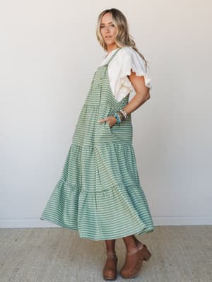 A green gingham tiered overall midi dress worn over a white ruffle-sleeve tee, paired with brown clogs and a stack of colorful bracelets — the kind of outfit that feels like a farmer's market and a good mood rolled into one.
