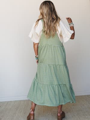 Shot from the back, this layered gingham midi dress in sage green is styled over a cream ruffle-sleeve top with brown platform clogs and a stack of beaded bracelets — casual and a little nostalgic, like a farmers market morning done right.