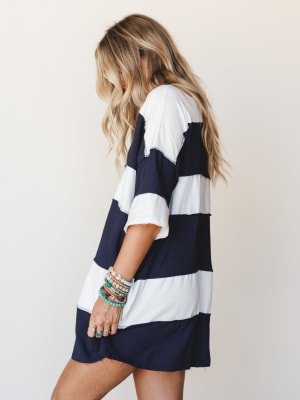 A side-profile shot of a model in an oversized navy and white wide-stripe dress with short sleeves, styled with a stack of beaded and stone bracelets — easy, lived-in, and effortlessly cool.