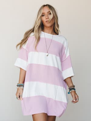 An oversized striped t-shirt mini dress in soft orchid pink and white, styled with stacked beaded bracelets and a delicate pendant necklace — the kind of effortless summer look that works as hard as you don't want to.