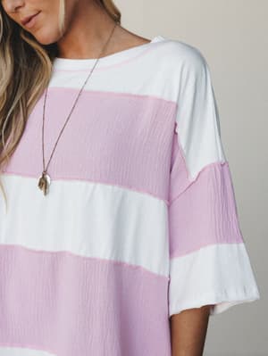 A close-up shot of a relaxed striped top or dress in orchid pink and white, styled with a delicate layered charm necklace — the kind of piece that makes a Saturday morning feel intentional.
