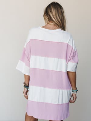 Shot from the back, a relaxed oversized striped dress in soft orchid pink and ivory reads effortlessly casual — styled with stacked beaded bracelets that feel more 'beach town summer' than anything.