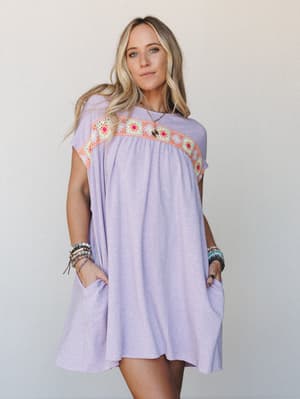 A relaxed lavender mini dress with a colorful granny square crochet yoke gives off that perfect 'grabbed something easy and still looked intentional' energy — styled with stacked bead bracelets and a pendant necklace for a lived-in boho feel.