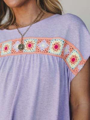 A close-up of a soft lavender knit top or dress with a cheerful granny square crochet panel across the chest — multicolor florals in peach, sky blue, yellow, magenta, and lilac — styled with layered gold chain necklaces for an easy, expressive everyd