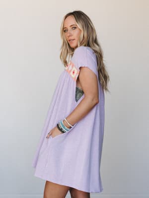 A lavender mini dress with a flowy, relaxed swing silhouette and a colorful granny square crochet panel at the chest — styled with stacked beaded bracelets for that effortless, feel-good summer energy.
