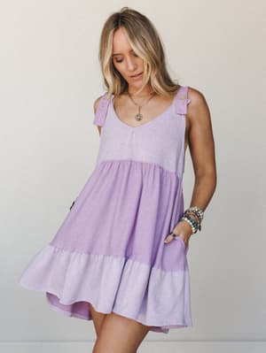 A breezy lavender babydoll mini dress with tiered ruffle hem and shoulder tie details, styled with layered gold necklaces and stacked boho bracelets — the kind of dress that makes a Tuesday feel like a vacation.