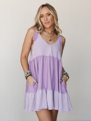 A lavender tiered babydoll dress with tie shoulders and side pockets, styled with layered gold necklaces and stacked turquoise bracelets against a soft neutral studio backdrop — easy, expressive, and made for warm-weather days.