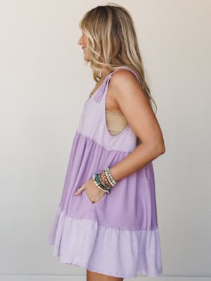 A tiered babydoll mini dress in soft lavender worn from the side, styled with stacked boho bracelets and a tan bandeau underneath — easy, warm-weather energy with a little personality built in.