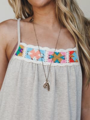 A close-up of a breezy spaghetti-strap dress in oatmeal knit, detailed with a multicolor granny square crochet panel across the chest — styled with a long brass feather charm necklace for that effortless, handmade-feeling boho.