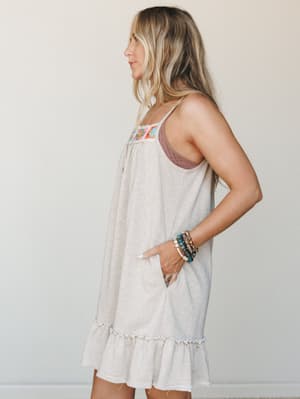 An airy spaghetti-strap mini dress in heathered oatmeal with a colorful crochet panel at the chest and a ruffled hem — the kind of dress you throw on and somehow look like you tried.