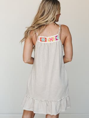 Shot from behind, a relaxed mini dress in heathered oatmeal shows off its standout crochet panel across the upper back — colorful granny squares in coral, pink, yellow, and turquoise edged in cream, with a ruffled hem that keeps the whole thing feeli