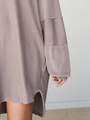 A close-up detail shot of an oversized mini dress in a soft mauve-taupe waffle knit, with paneled long sleeves and a curved hem — the kind of piece that looks like it came from your coziest daydream.