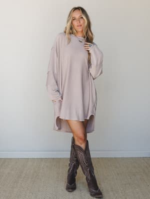 An oversized ribbed mini dress in a soft oat-mauve tone styled with dark brown cowboy boots — the kind of effortless outfit that looks like you tried just enough.