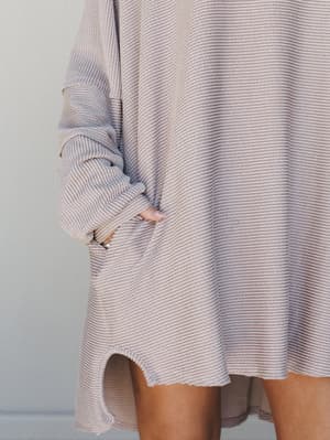 A close-up detail shot of an oversized ribbed mini dress in a soft oat tone, hand tucked into a front pocket with a long sleeve casually pushed up — the kind of dress that looks like you tried just enough.