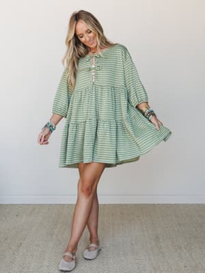 A breezy green gingham mini dress with tiered ruffles and a playful tie neckline, styled with stacked turquoise bracelets and woven flats — the kind of dress that makes you want to twirl in a farmers market.