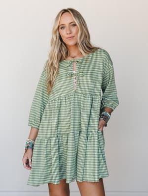 A breezy green gingham mini dress with a tiered babydoll silhouette and lace-up front detail, styled with stacked boho bracelets against a clean neutral backdrop — the kind of dress that makes a farmers market run feel like an occasion.