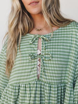 A close-up of a green gingham mini dress with a charming lace-up front detail and crochet trim — the kind of piece that makes a Tuesday feel like a picnic.
