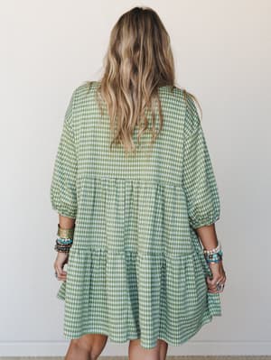 A breezy green gingham mini dress shot from the back, with tiered ruffles and billowy sleeves — styled with stacks of colorful bracelets that give it a relaxed, personal feel.