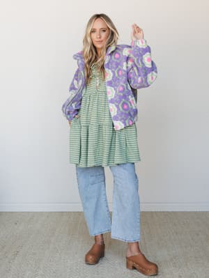 A layered outfit full of playful pattern-mixing — a green gingham tiered mini dress styled under a floral sherpa jacket, worn over wide-leg light wash jeans with brown wooden clogs, shot against a clean studio wall with an effortlessly cool, free-spi
