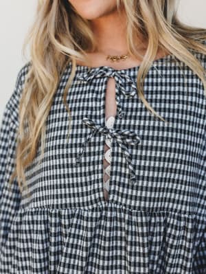 A close-up detail shot of a black and white gingham mini dress with a sweet self-tie keyhole neckline and crochet button placket — the kind of piece that feels equally at home at a farmers market or a rooftop brunch.