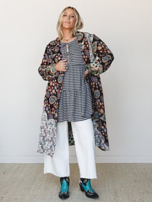 A layered boho outfit built around a gingham tiered mini dress worn under a floor-length paisley and floral patchwork duster, grounded by white wide-leg pants and turquoise cowboy boots — the kind of mix-and-match energy that looks effortless but is