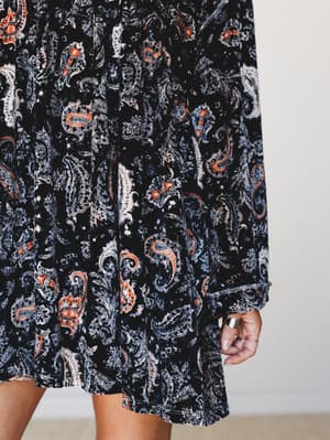 A close-up detail shot of a flowing velvet mini dress in a rich black paisley print with rust orange and dusty blue accents — the kind of dress that looks like it has a whole personality before you even put it on.