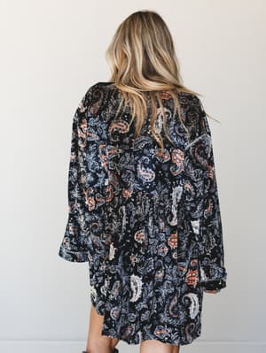 Shot from behind, a model in a flowing velvet mini dress covered in a bold black-and-rust paisley print — the kind of dress that feels as good as it looks, worn with effortless loose waves and a casual, lived-in confidence.