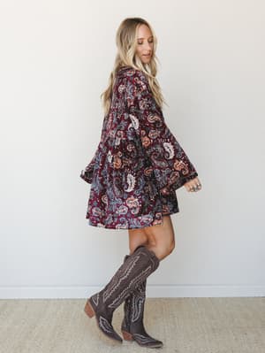 A flowy velvet paisley mini dress in deep burgundy paired with tall brown cowboy boots — the kind of outfit that makes a Tuesday feel like a road trip.