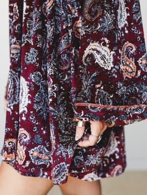 A close-up detail shot of a rich burgundy velvet mini dress in an all-over paisley print, hand peeking through a wide bell sleeve with stacked gold rings — the kind of dress that feels like fall itself.