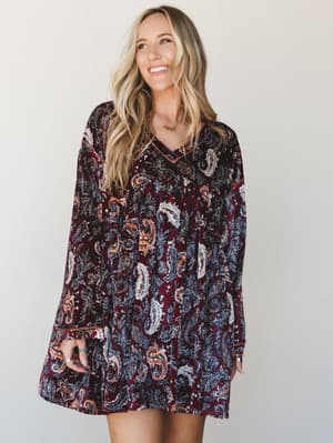 A smiling model in a flowy velvet mini dress covered in a rich burgundy paisley print — the kind of dress that looks equally at home at a fall farmers market or a candlelit dinner.