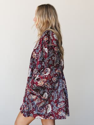 A side profile shot of a paisley mini dress in deep burgundy with slate blue, cream, and peach accents — the kind of print that looks like it belongs at a fall concert or a weekend somewhere with good wine.