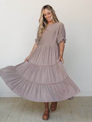 A smiling model twirls in a dusty mauve ribbed maxi dress with a tiered skirt and ruffled sleeves, styled with tan clogs against a clean white studio wall — the kind of dress that makes you want to spin.
