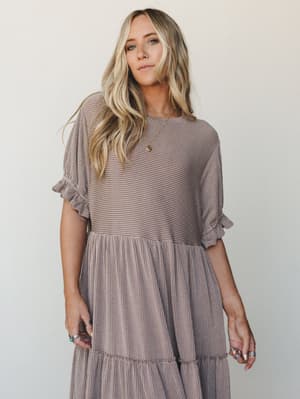 A tiered maxi dress in warm acorn brown with a subtle stripe texture, styled with layered gold and turquoise jewelry — the kind of dress that feels effortless whether you're running errands or staying for dessert.