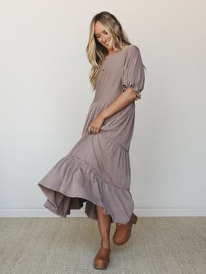 A flowy striped maxi dress in warm mauve tones moves mid-twirl on a smiling model — the tiered ruffle hem and puff sleeves give it that effortless, lived-in feel, grounded by tan clogs and a turquoise ring.
