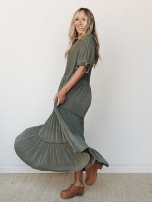 A model mid-twirl in a dark olive tiered maxi dress with subtle ribbed texture and flutter sleeves, paired with tan studded clogs against a clean white studio wall — the kind of dress that makes you want to spin just to see what it does.
