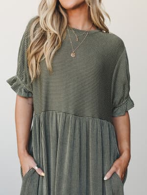 A close-up torso shot of a textured striped maxi dress in dark olive, with ruffle sleeve cuffs, a gathered empire waist, and deep pockets — the kind of dress that looks like you tried just the right amount.