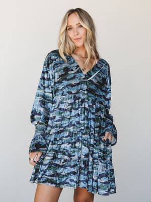 A flowy blue camo velvet mini dress with tiered skirt and smocked cuffs, styled with layered gold necklaces against a clean studio backdrop — the kind of dress that makes a statement without trying too hard.