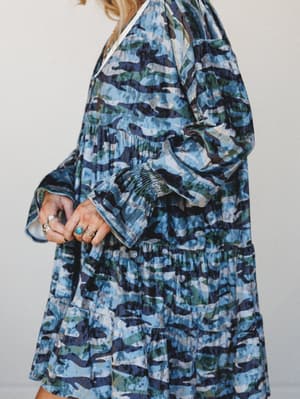 A close-up side view of a tiered mini dress in a blue-toned camo print, with smocked cuffs and long sleeves — worn with a stack of silver and turquoise rings that give it that effortless, collected feel.
