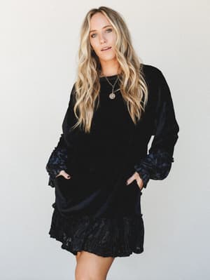 A relaxed mini dress in deep black velvet with contrast ruffle trim at the hem and sleeves, styled with a gold pendant necklace — the kind of dress that works just as hard at a holiday party as it does with boots on a Tuesday.