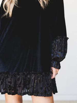 A close-cropped detail shot of a black velvet mini dress with a flocked floral ruffle hem and matching gathered cuffs — moody and romantic with just the right amount of drama.